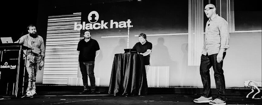 BT6 presenters on the Black Hat stage, with a robot dog beside the presentation area.